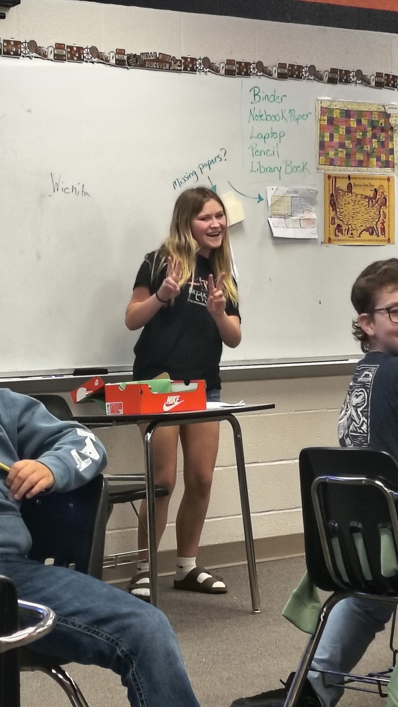 Students seated at desks in a classroom, one girl stands at a table with items, speaking.