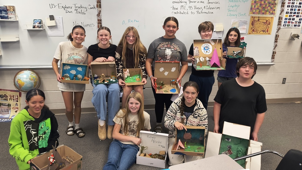 Students in a classroom pose with cardboard boxes holding dioramas of various scenes, some standing, some sitting, against a whiteboard background.