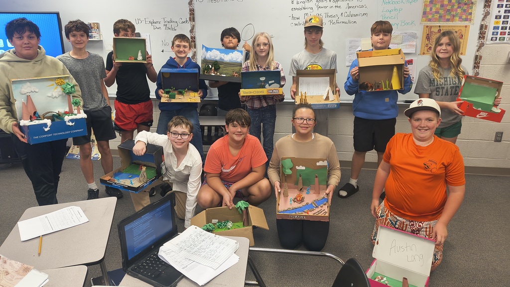 Nine children hold up cardboard boxes in a classroom. Some boxes contain a model landscape. A whiteboard and posters are behind them.