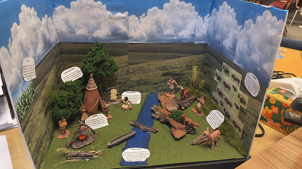 Miniature diorama shows a river with trees and a village. Small figurines depict people. White labels attached to objects.