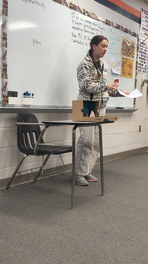 A student is standing at a table with a box on it, holding a paper, and speaking.