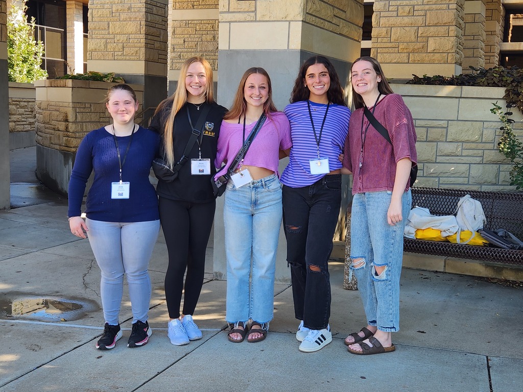Five girls in casual clothes stand on a sidewalk, wearing lanyards and holding IDs, in front of a building.