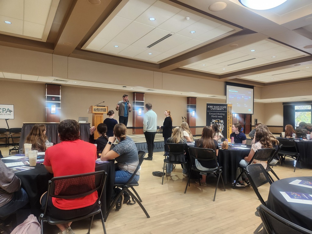 People seated at tables, facing a stage with a person standing, another seated, and a projector screen.