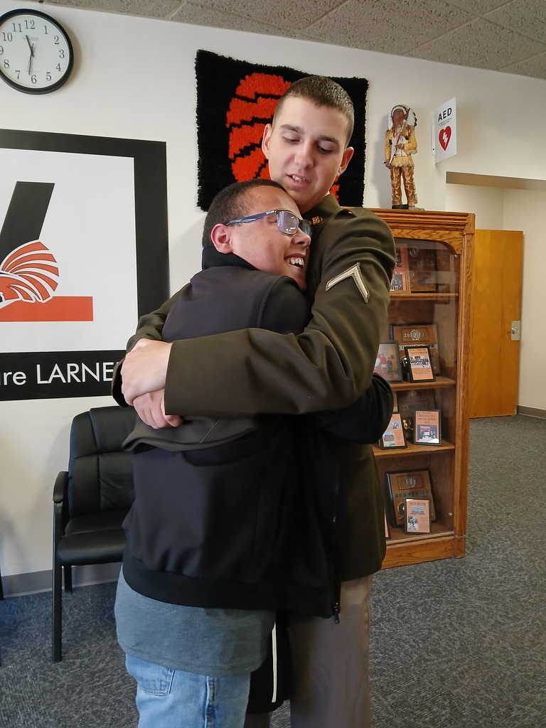 Two people hug in a school room; a man in a military uniform. Behind them is a clock, a bulletin board, and a cabinet.