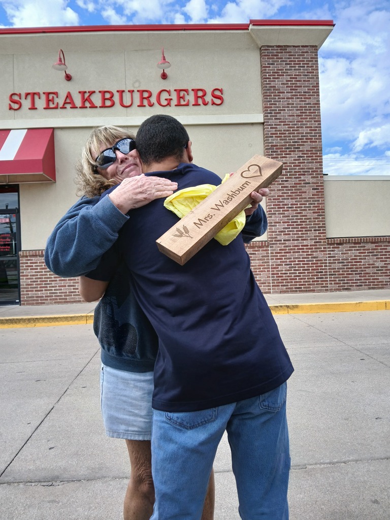 Adult holds a box while hugging another adult in front of a restaurant with a red awning.