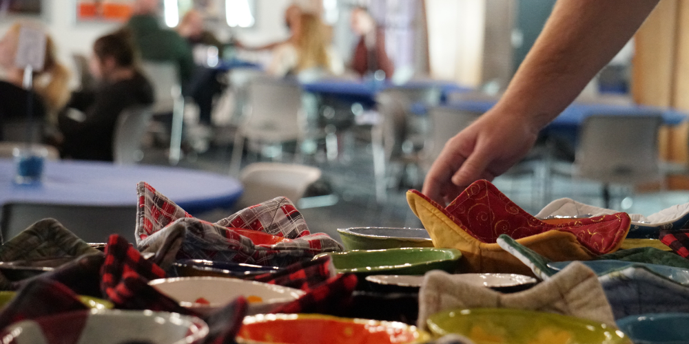 A hand reaches toward a table covered in colorful ceramic bowls and bowl cozies as guests share a meal in the background