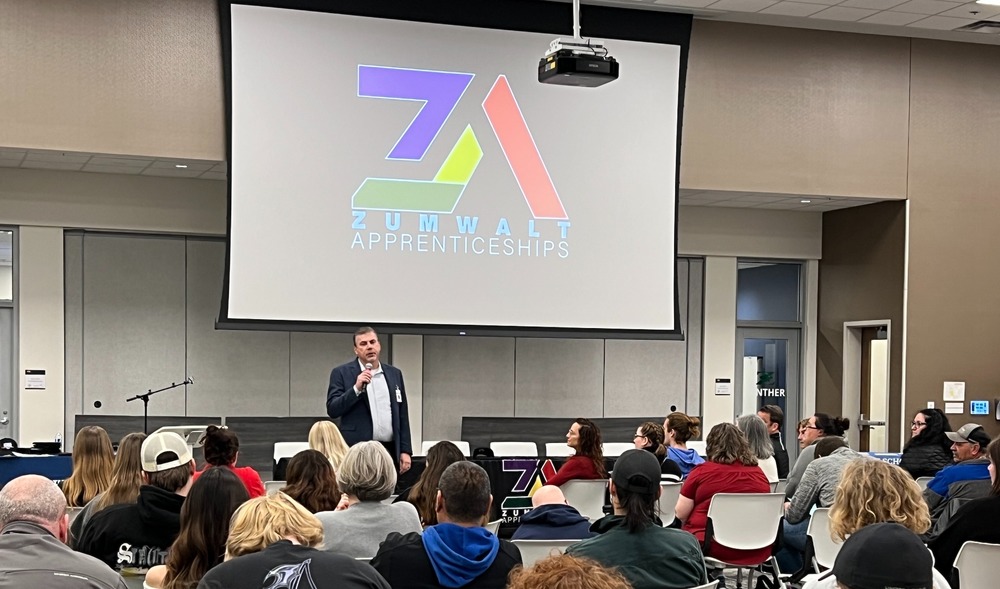 Program director addresses large crowd in front of screen featuring Zumwalt Apprentice logo