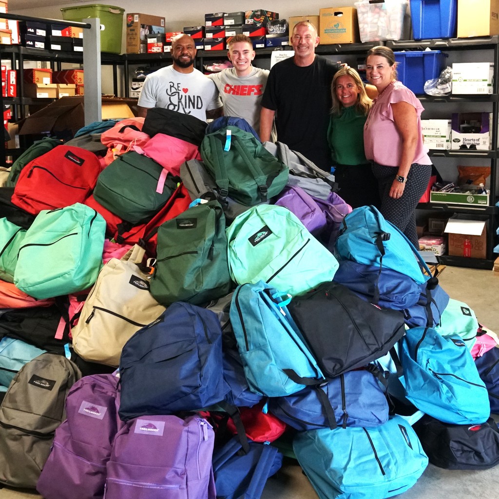 Donors and staff stand arm-in-arm behind a mountain of 300 donated backpacks stuffed with school supplies