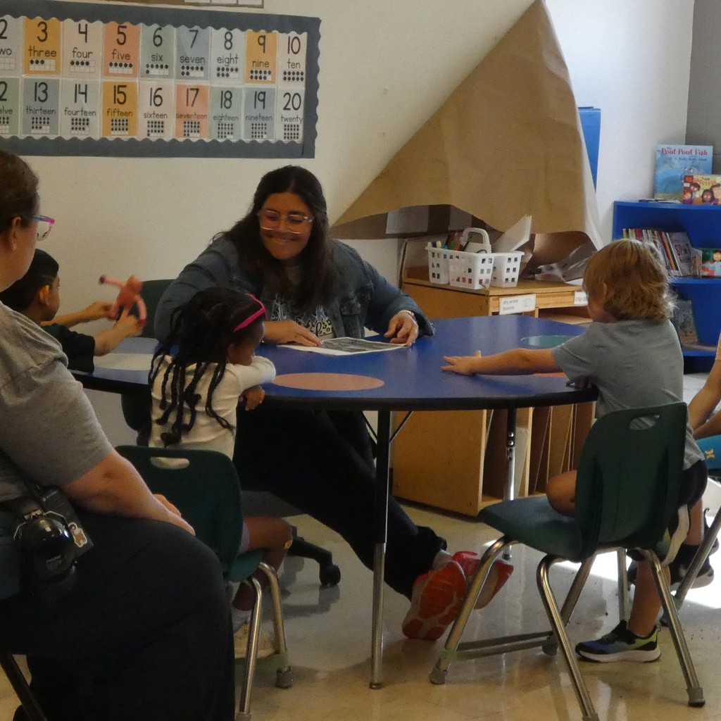 Students surround the teacher at a half-circle table as she smiles and learns about each student's summer