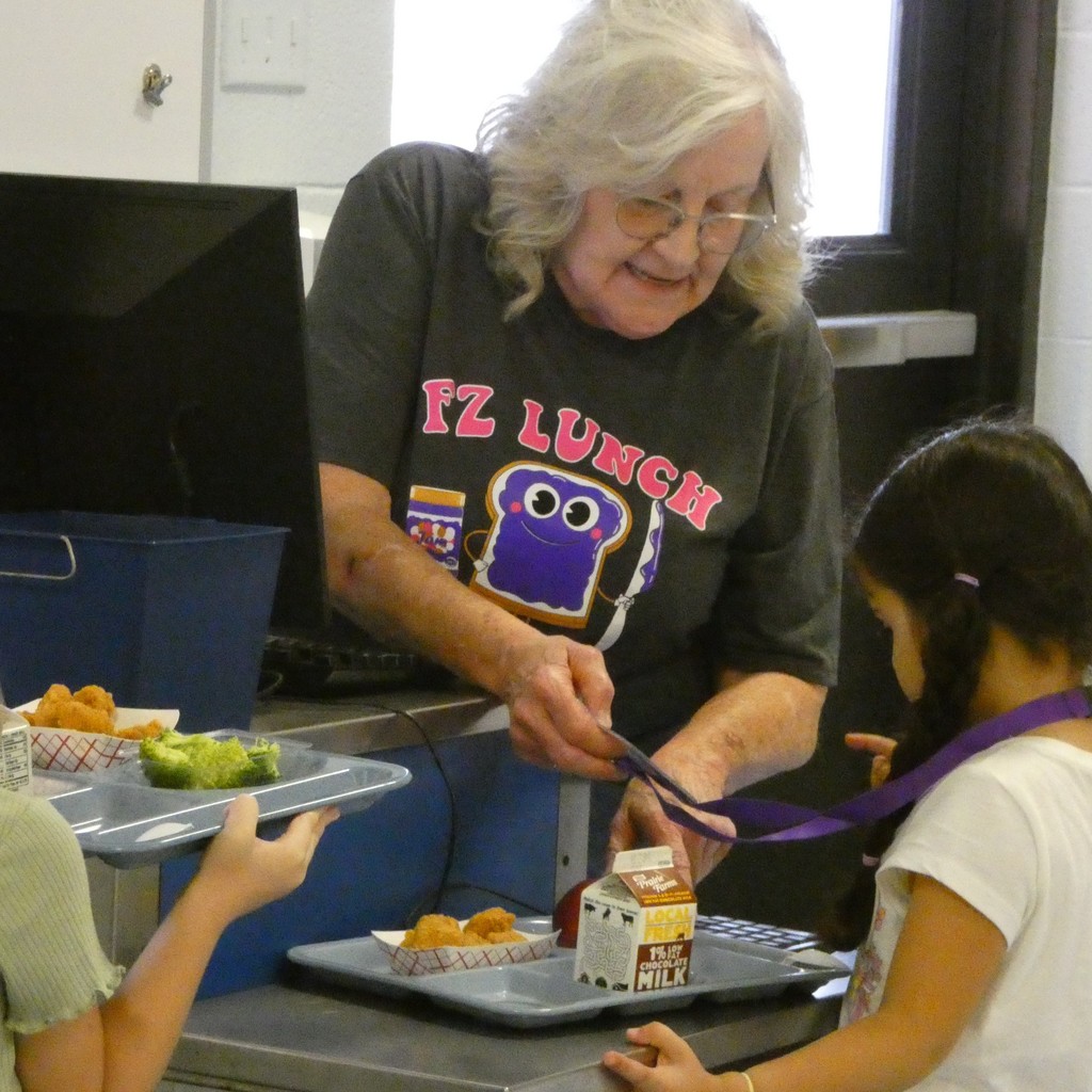 A cafeteria cashier leans in to help a kindergartener complete her purchaces of nuggets, and apple and chocolate milke