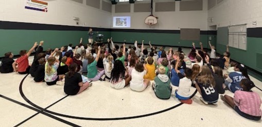 Students in rows on the gym floor shove their hands in the air to ask questions of the playground designer during presentation