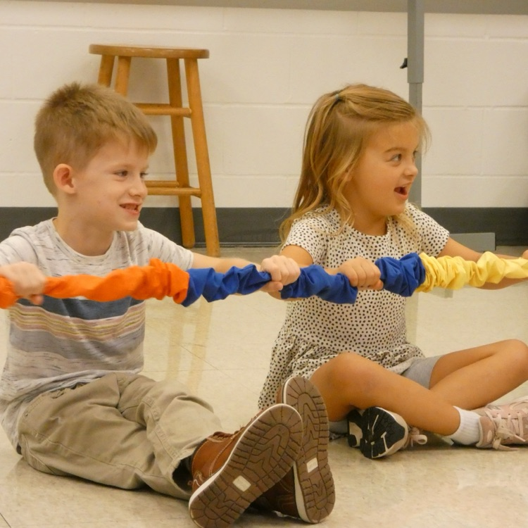 Students sit on the floor with the colorful stretchy band, smiling and laughing as they sing and follow the teacher's motions 