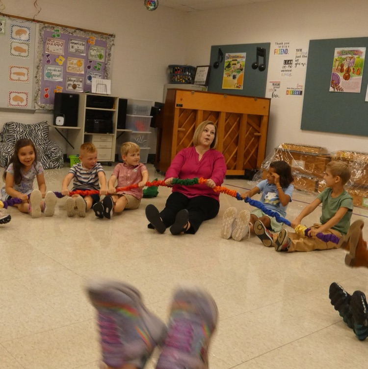The teacher on the floor in a circle of students holding a colorful stretchy band and leaning back as far as they can go without laying down