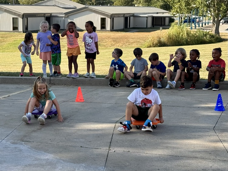 Two students racing on scooters while their class cheers them on  