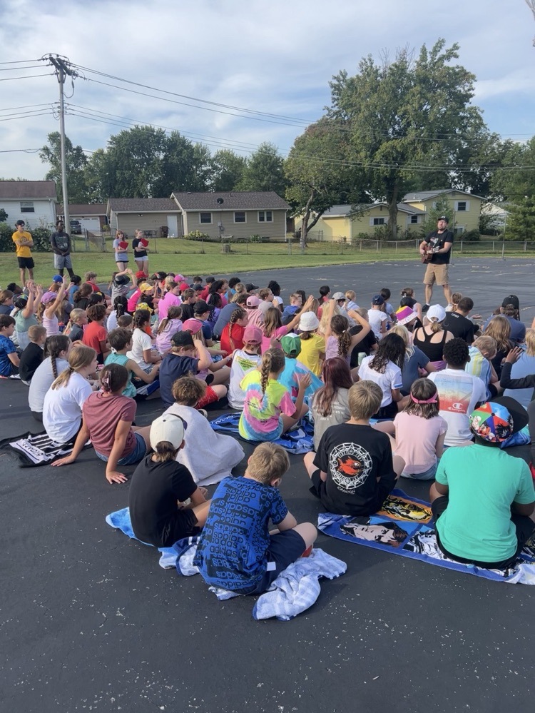 camp songs time! students sitting on concrete with a man playing a guitar