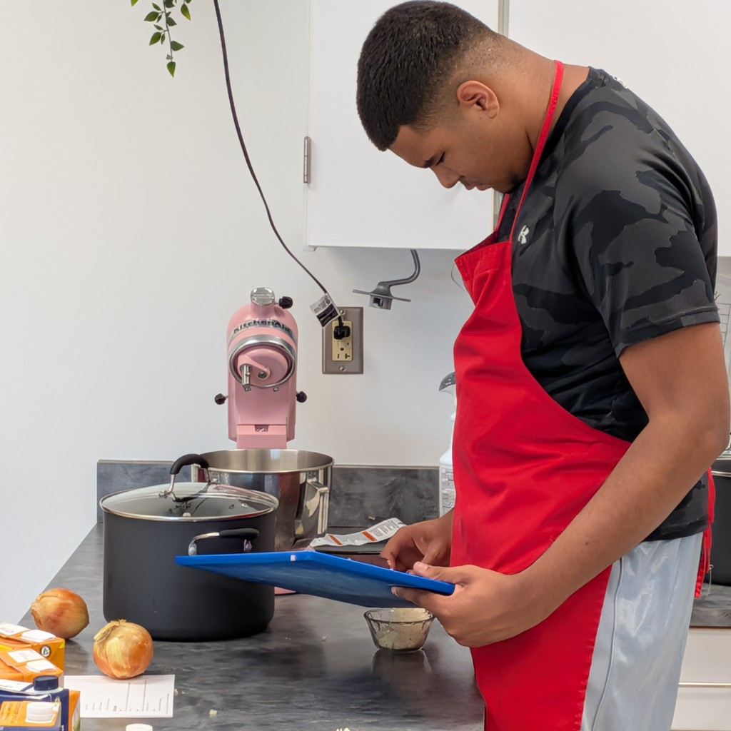 a student in a red apron checks the recipe as he carefully measures.