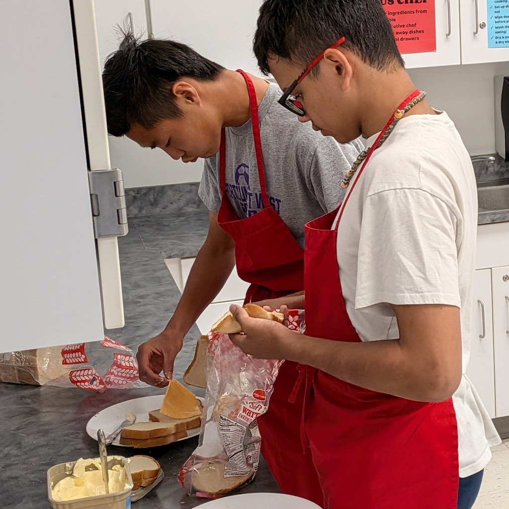 A pair of students in red aprons assembles cheese sandwiches to be grilled