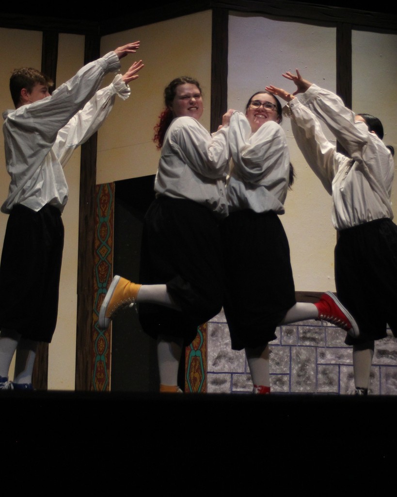 Two student actors strike a "foot pop" pose under an arch that is built by other actors raising their arms