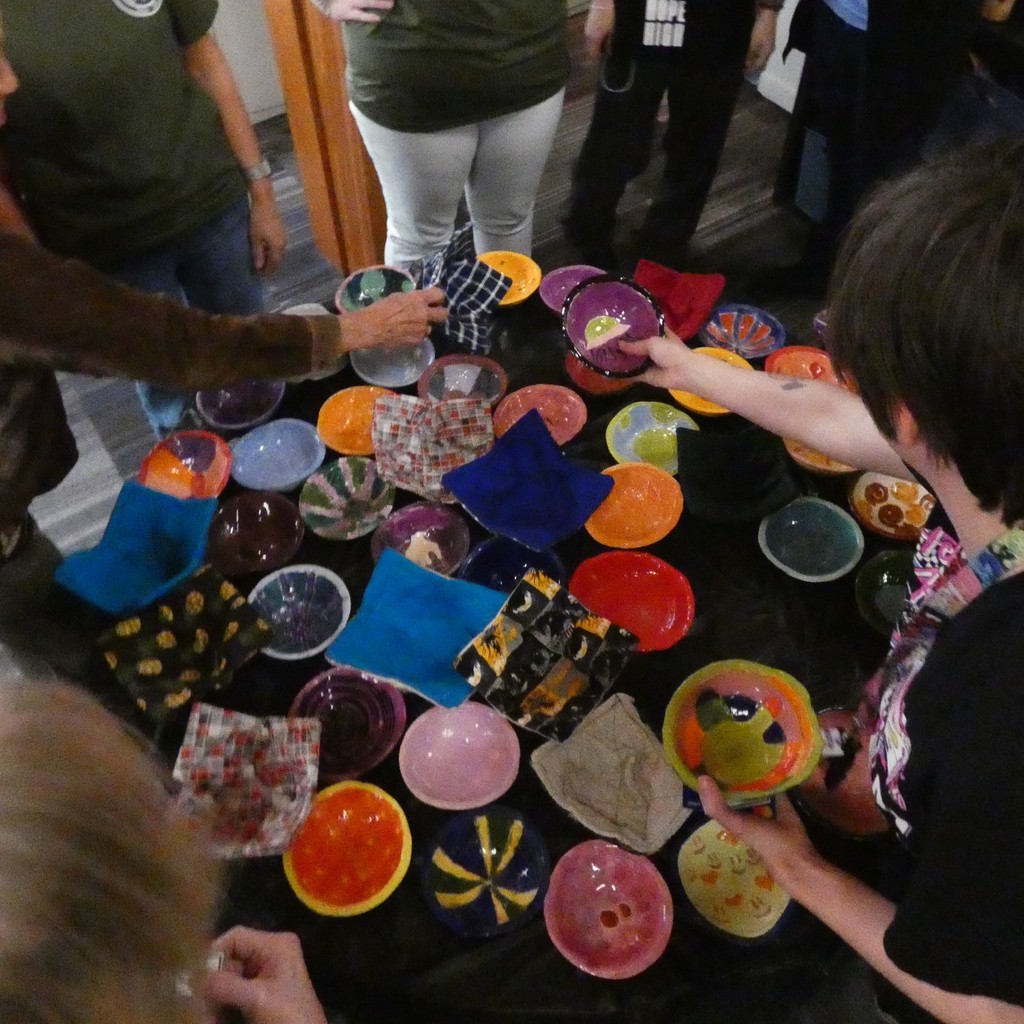 A circular table loaded with colorful bowls and bowl cozies is surounded by guests making their selections