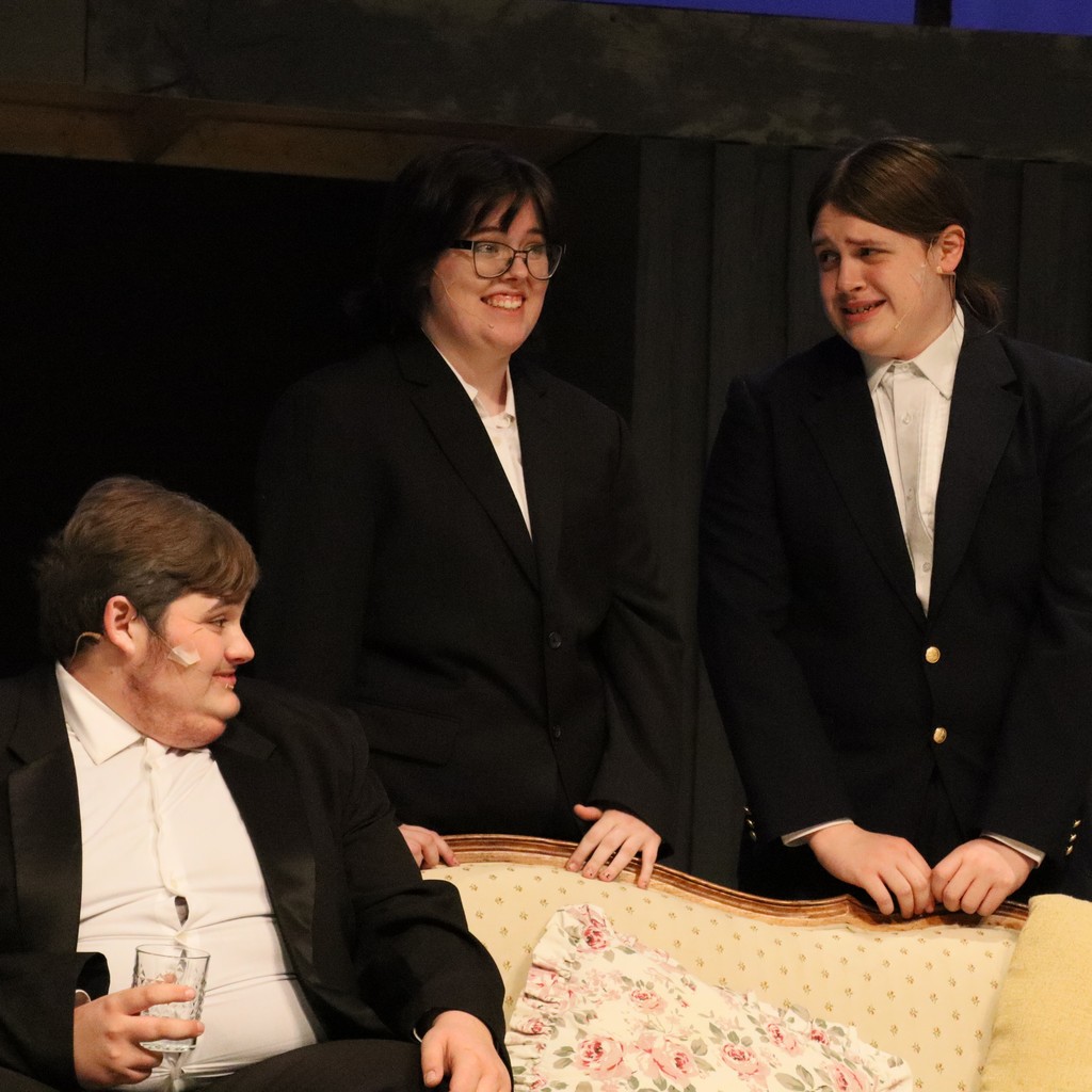 Three student actors in black suits with white shirts laugh during a scene