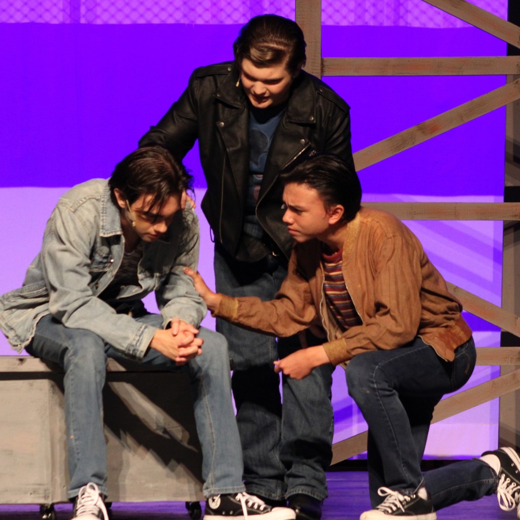 Student actors with slicked back hair perform a scene, two consoling the third 