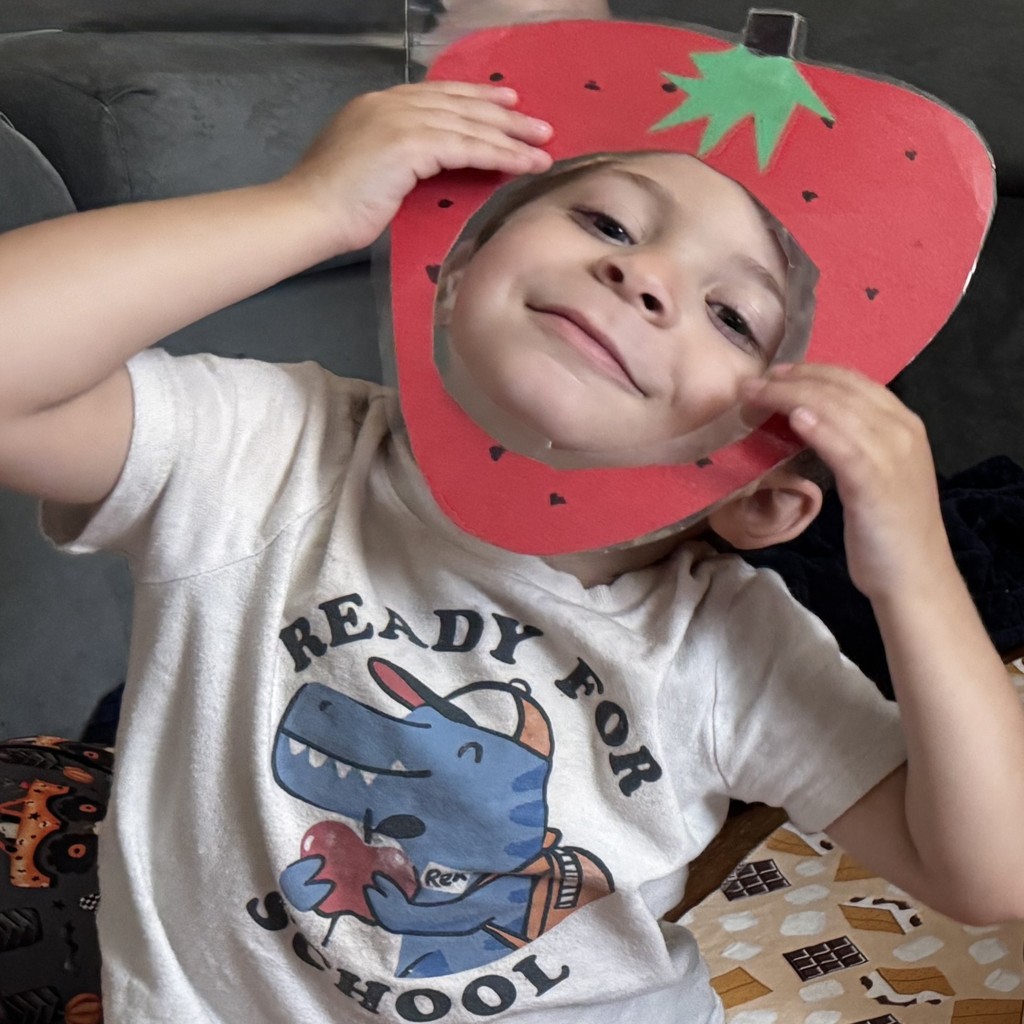 Preschooler in a T-shirt featuring a cartoon dino wearing a backpack and the words "Ready for School" grins as he sticks his face through a paper strawberry frame