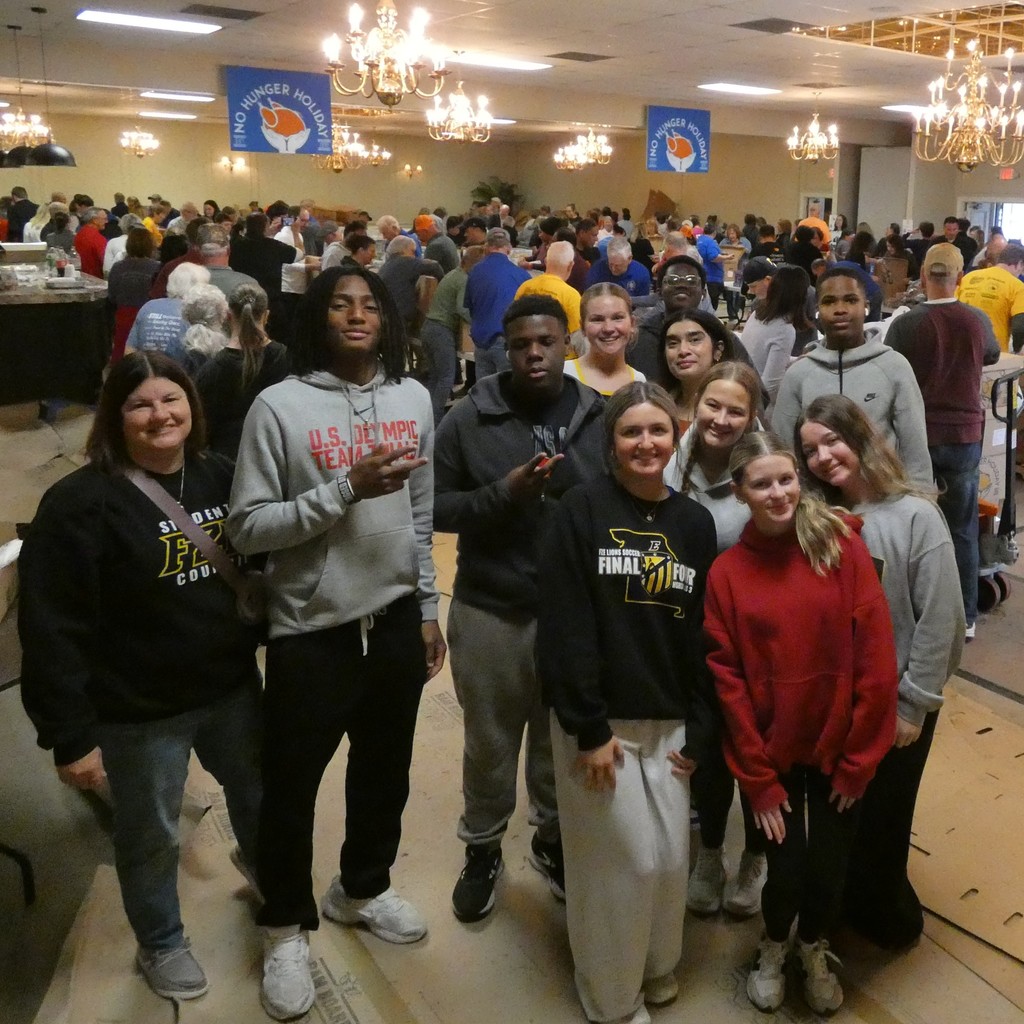Students pause packing meals for a photo, with a ballroom crowded with volunteers in the background