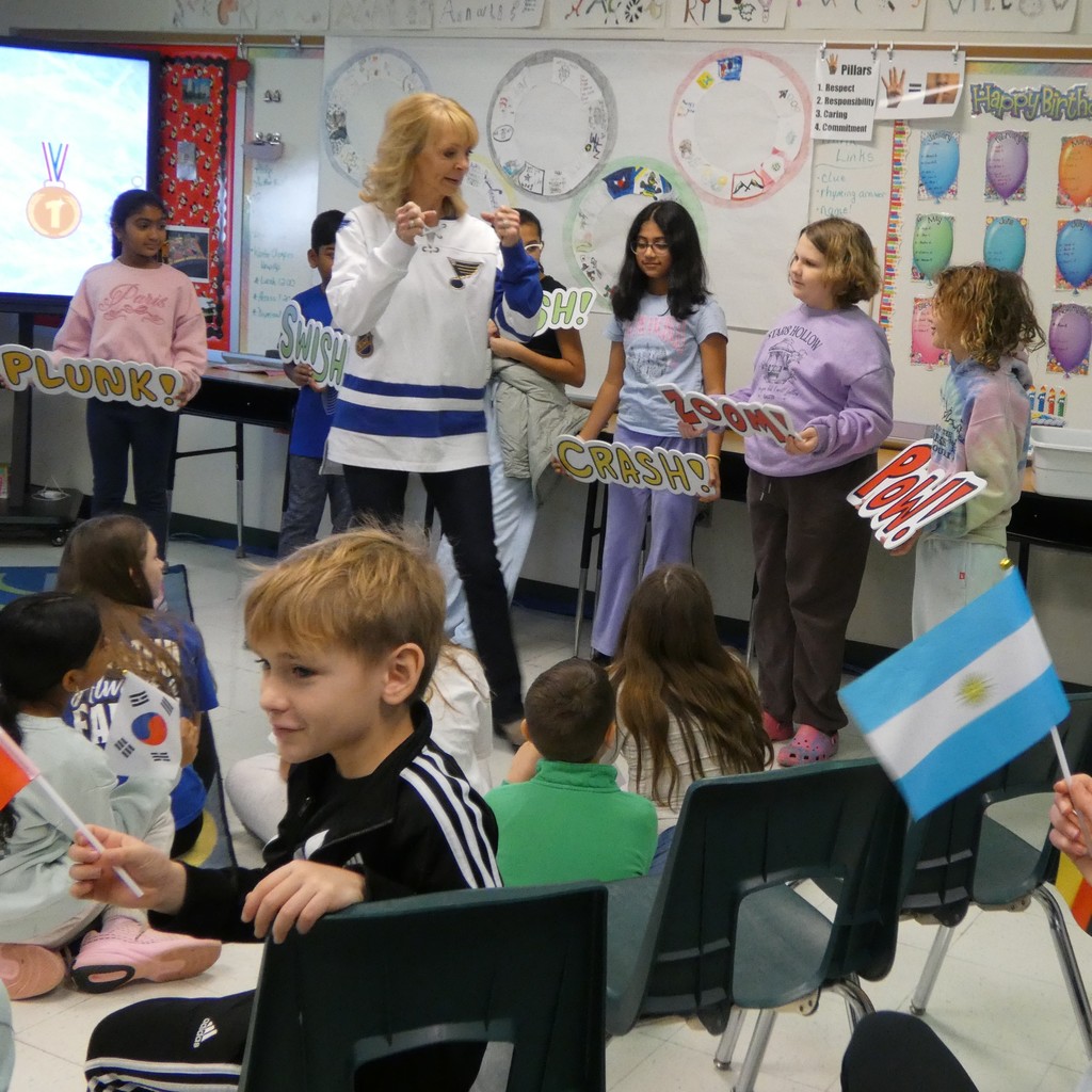 seated students wave miniature flags from different countries while guest speaker coaches Sound Engineers in charge of acting out the words on signs they hold such as Pow!!
