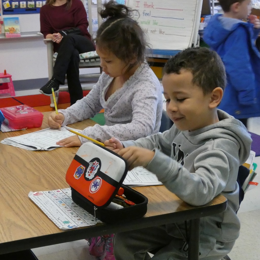 Kindergarteners get to work, sitting at the table with pencil boxes and worksheets