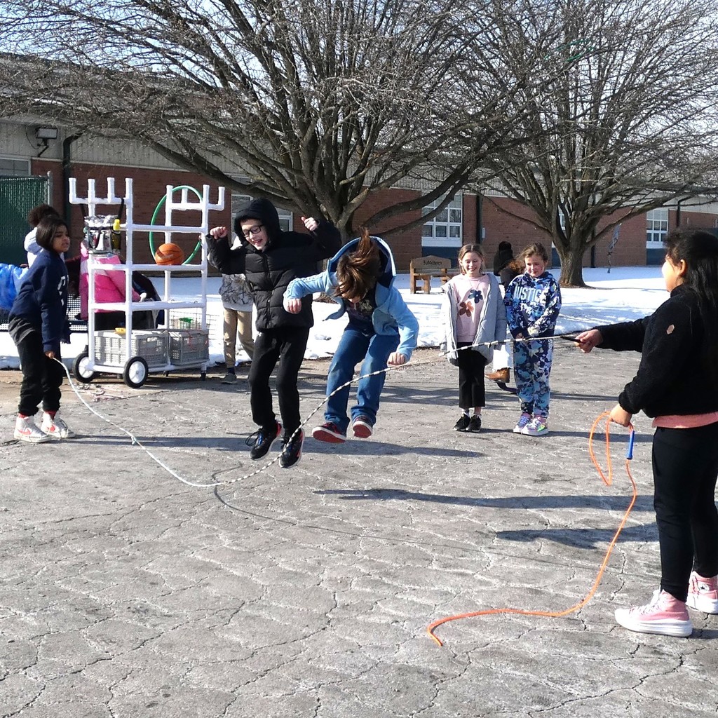 students on dry concrete jumping rope. Two turn the rope while laughing, two jump while laughing, two smile as they wait their turn