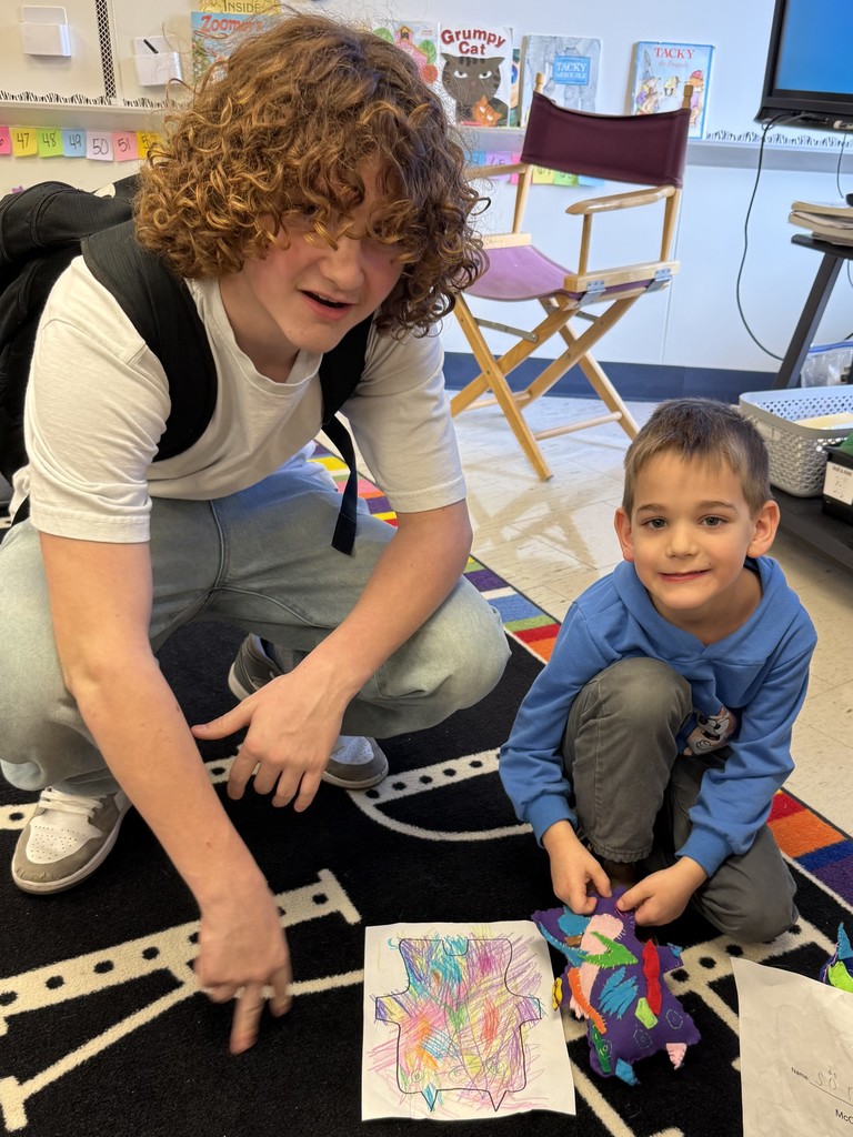 An 8th grader crouches next to a smiling kindergartener who compares his monster with his illustration