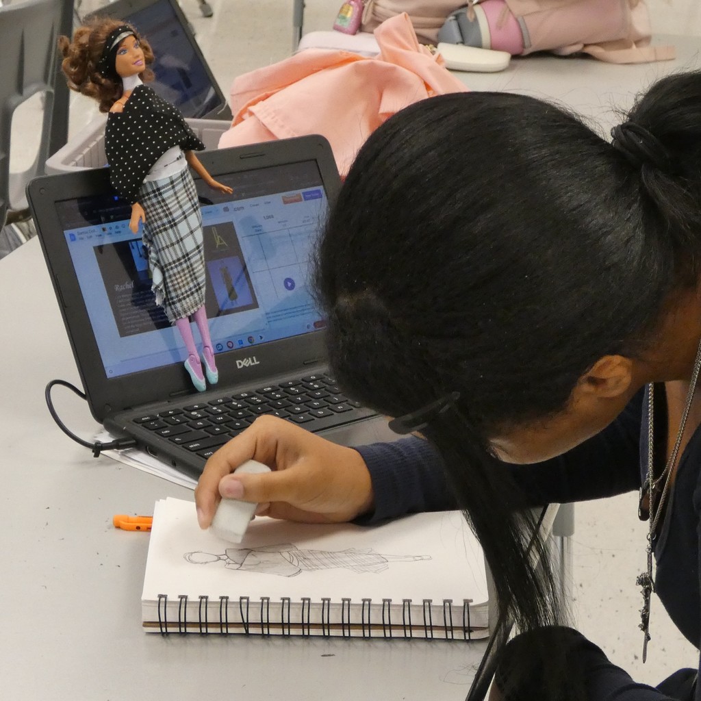 A Barbie doll wrapped in fabric is perched on Chromebook as fashion student sketches the design it inspires