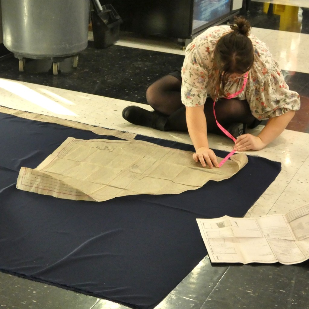 student with a flexible pink tape measure around neck works on the floor to place pattern on fabric 