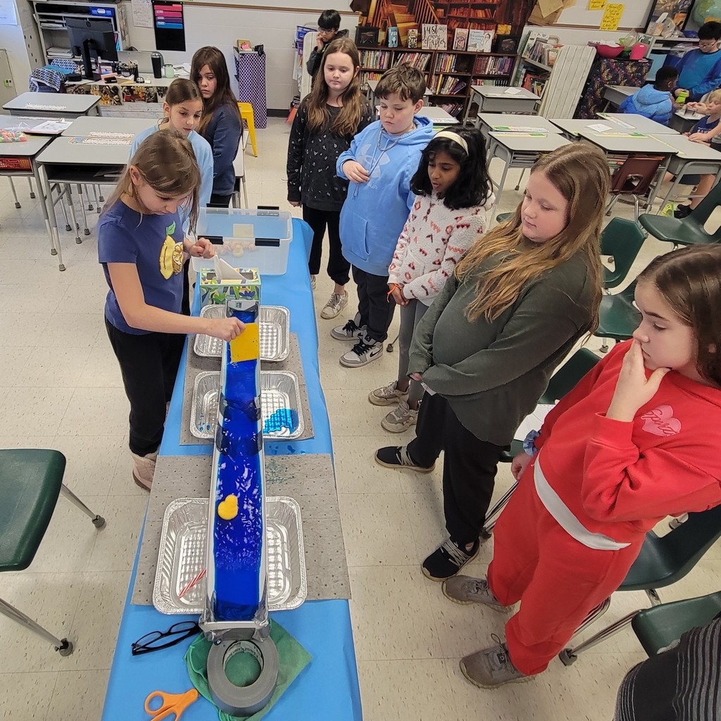 A student drags a plastic scraper through dyed water in a trough, creating wave action for a rubber duck to ride