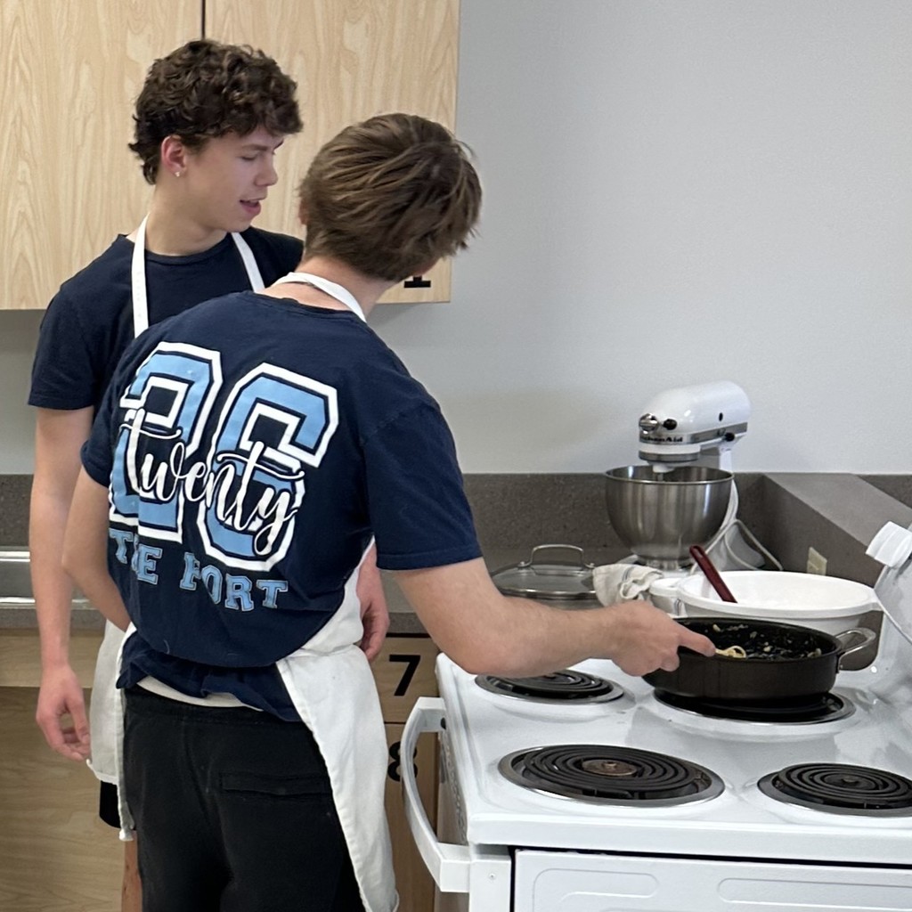 Students in Senior Year T-shirts and white aprons tend to a pot on a stove with heating coils