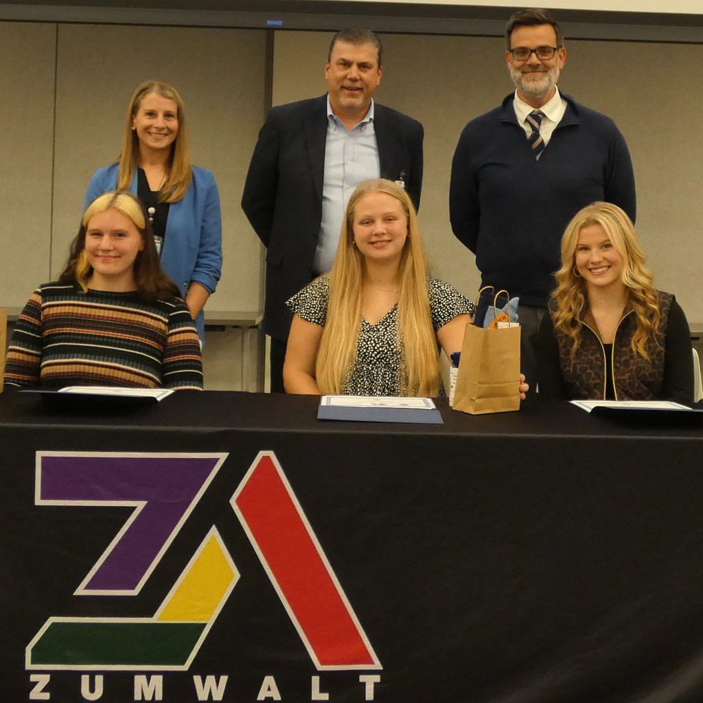 Three students sit at a table behind the Zumwalt Apprentice logo, flanked by proud teachers