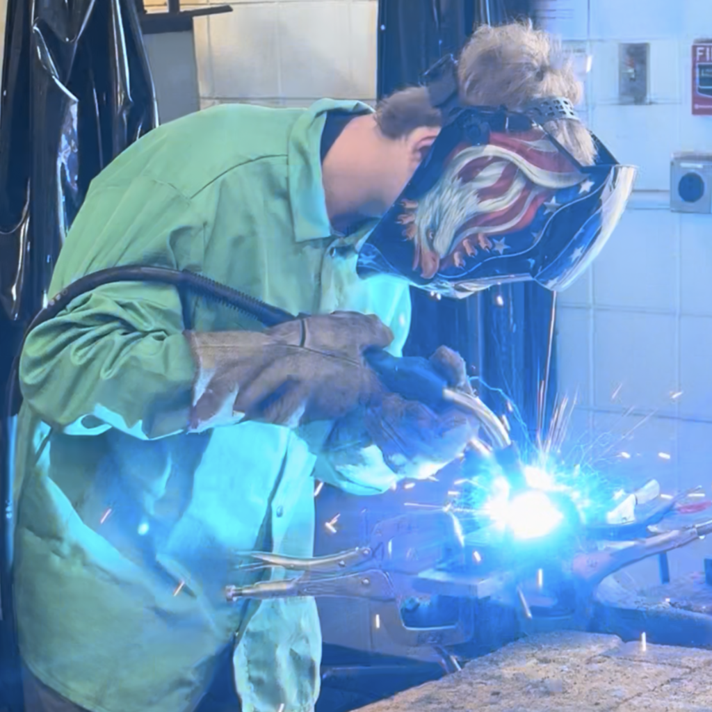 Sparks fly as a student in a protective helmet with patriotic design welds to components together