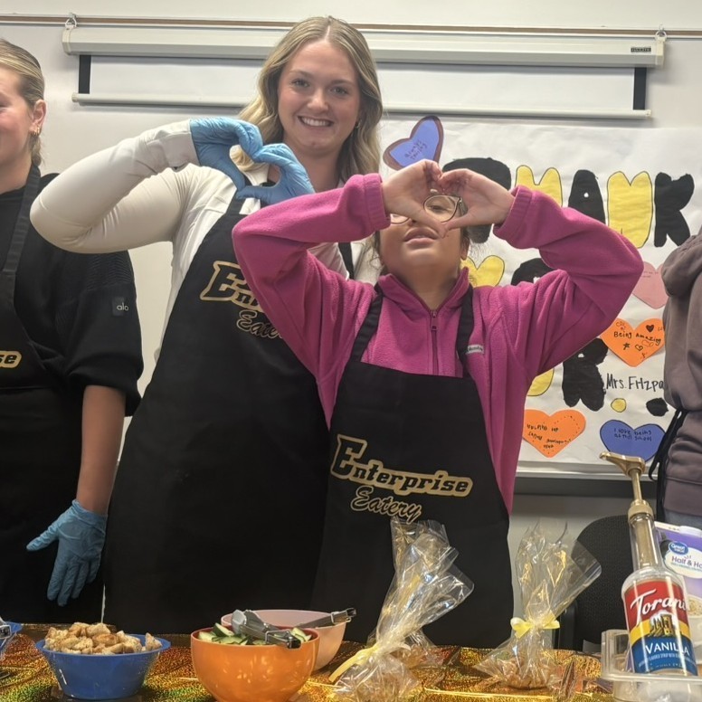 Between customers, two students in black aprons make heart hands for the camera