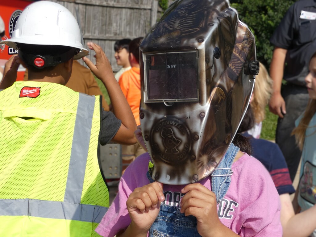 student in pink tshirt and overalls tries on welding helmet while classmate models safety vest and hard hat