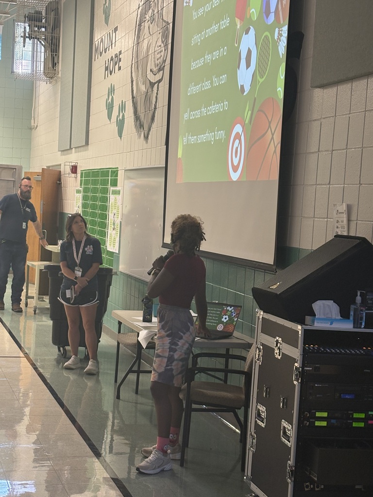 Three adults are presenting to students in a cafeteria.