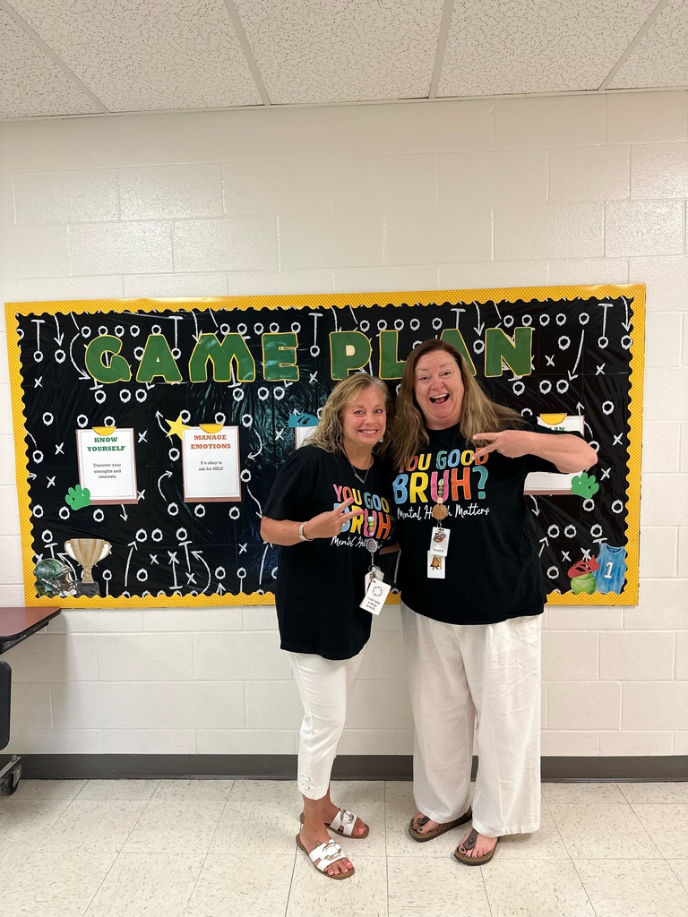 Two teachers making peace signs and posing for a picture in front of a bulletin board that says GAME PLAN on it.