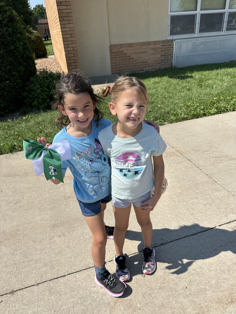 Two girls are standing outside the school building. One has a hair bow in her hand that has a Mount Hope Husky on it.