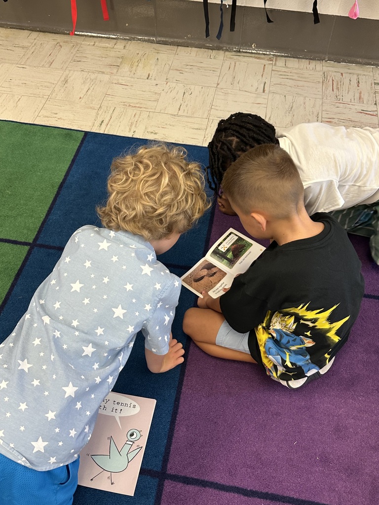 Three boys are reading a book together on a rainbow colored carpet.
