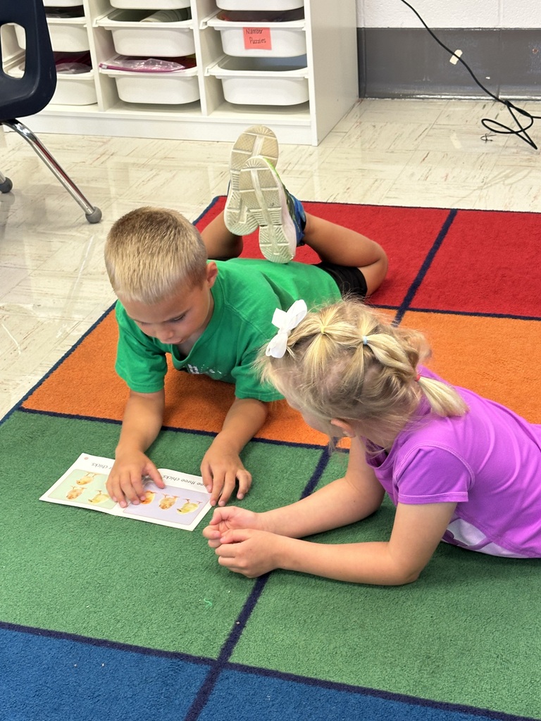 Two students reading a book together. One is a boy and one is a girl. They are on a rainbow colored rug.