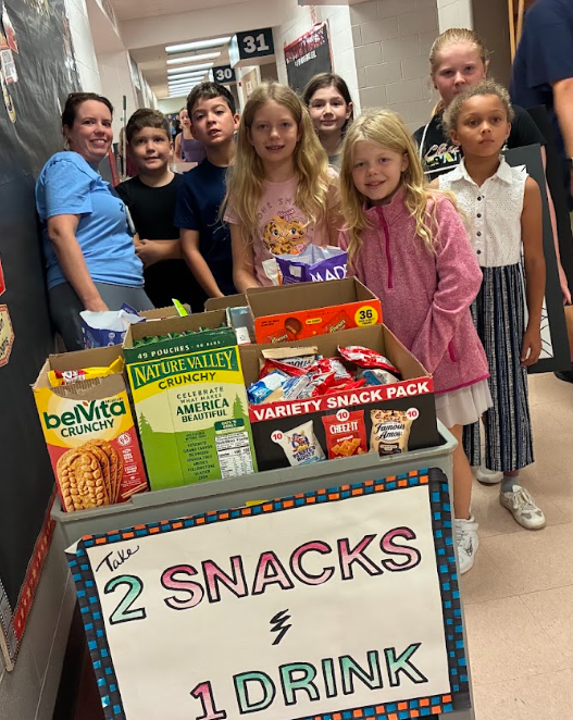 A teacher and a group of elementary students stand behind a rolling cart filled with boxed snacks, featuring a sign that reads "Take 2 SNACKS & 1 DRINK."