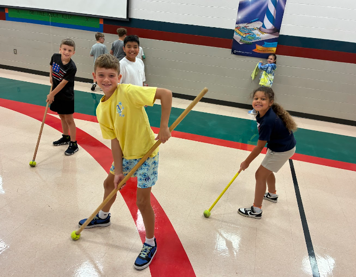 Elementary school students in a gymnasium smiling at the camera while using long wooden handles tipped with tennis balls to push or sweep items along the floor.