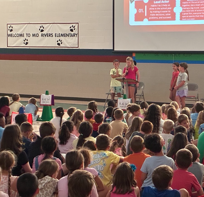 A young girl speaks into a microphone at a podium during a school assembly in a gymnasium full of seated students, with a banner reading "Welcome to Mid Rivers Elementary."