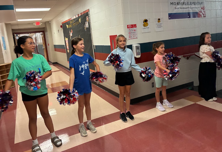 Five elementary school students lined up in a school hallway holding blue and red cheer pom-poms.