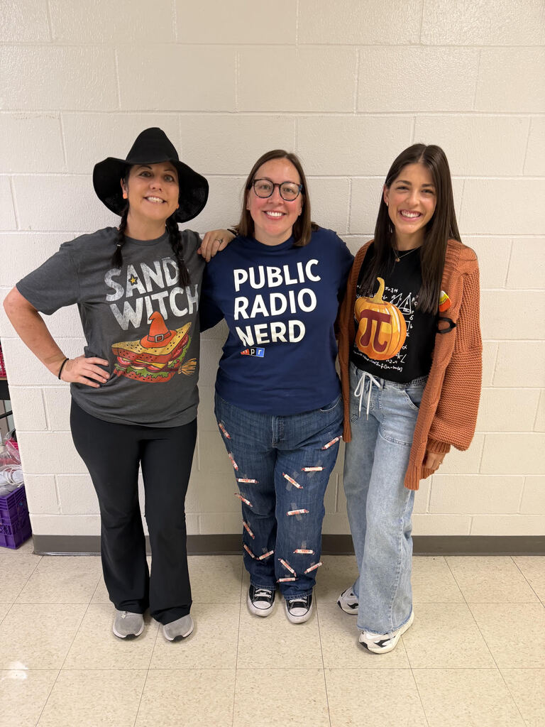 Three teachers standing next to each other. One is wearing a shirt that says sandwitch. Alnother is wearing a shirt that says public radio nerd. A third is wearing a shirt with a pumpkin and the pi symbol on it. They are supposed to be a sandwitch, smarty pants, and pumpkin pi.