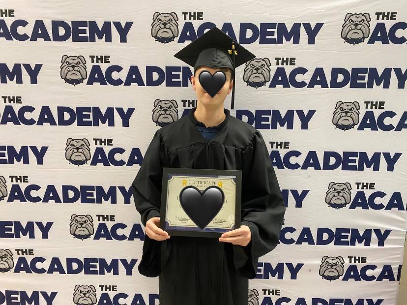 A graduate stands holding a certificate in cap and gown. Back drop says The Academy and has a  bulldog on it.